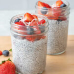 Two single serving jars of chia pudding topped with fresh berries and coconut shreds, surrounded by scattered fruit.