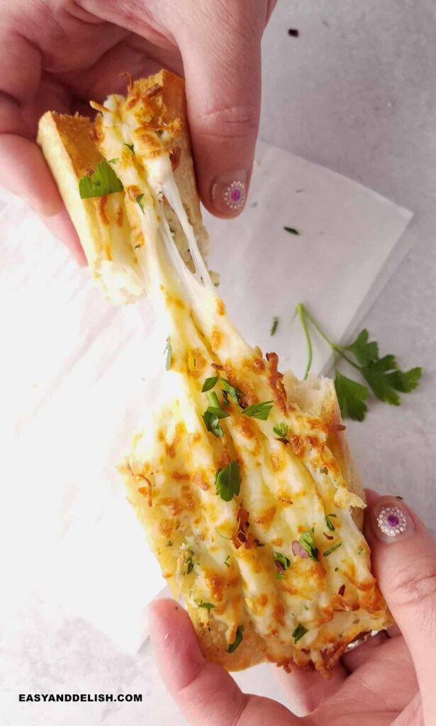 Hands pulling a cheesy garlic bread apart to show the melty cheese between the pieces. 