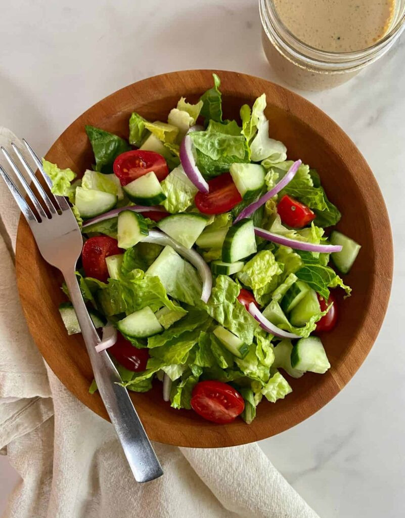 Top view of a fork resting on the side of a wooden salad bowl with a house salad served in it and dressing in a mason jar on the side. 