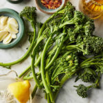 Fresh broccolini, lemons, garlic, red pepper flakes, olive oil, and salt on marble countertop