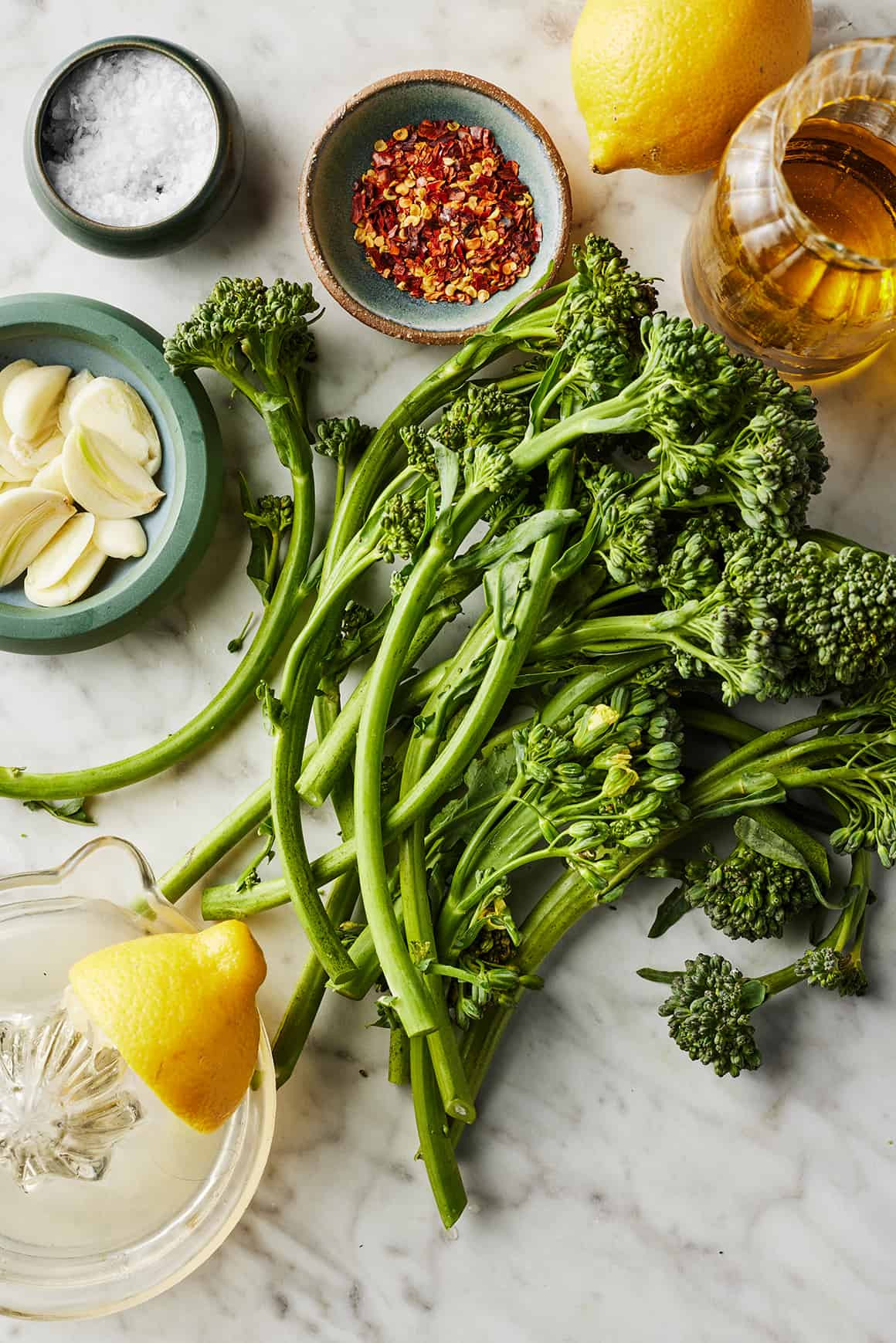 Fresh broccolini, lemons, garlic, red pepper flakes, olive oil, and salt on marble countertop