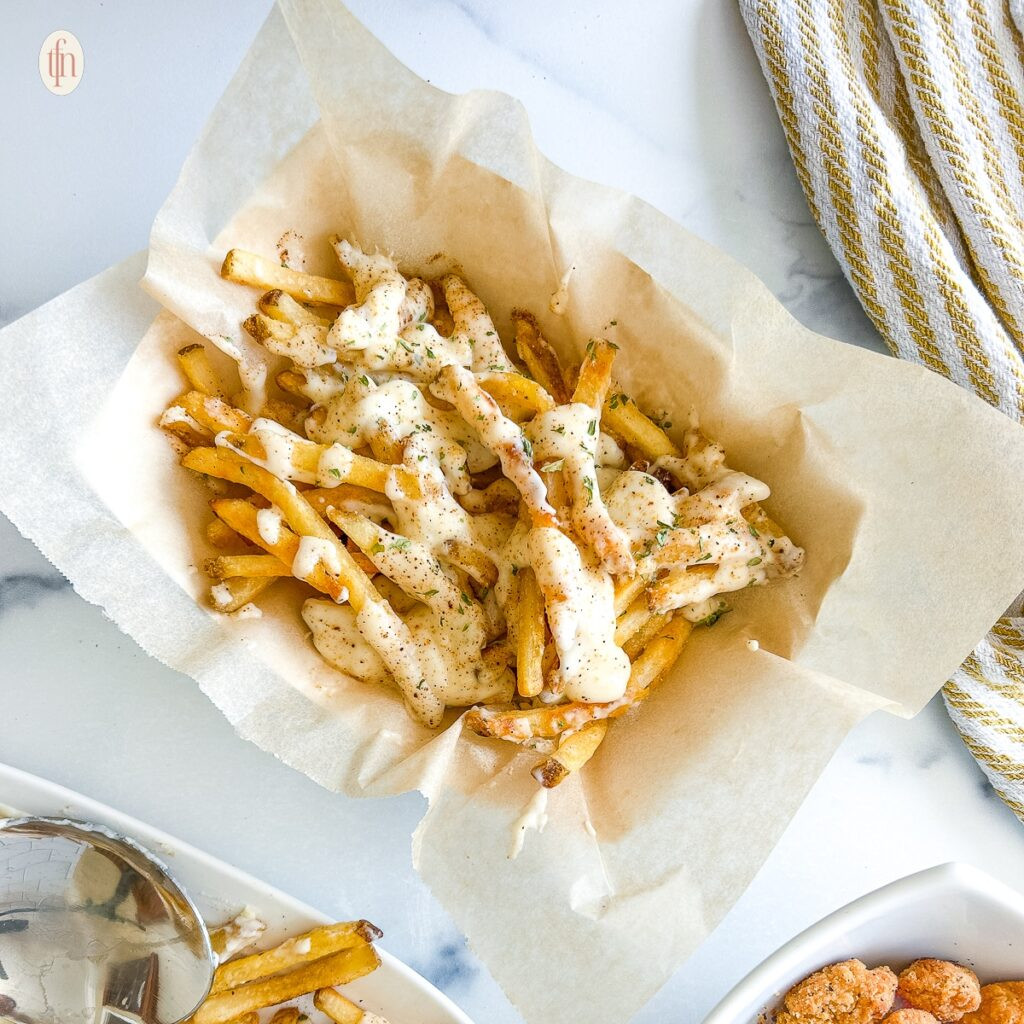 Louisiana Voodoo Fries topped with a creamy white sauce in a parchment paper covered dish. 